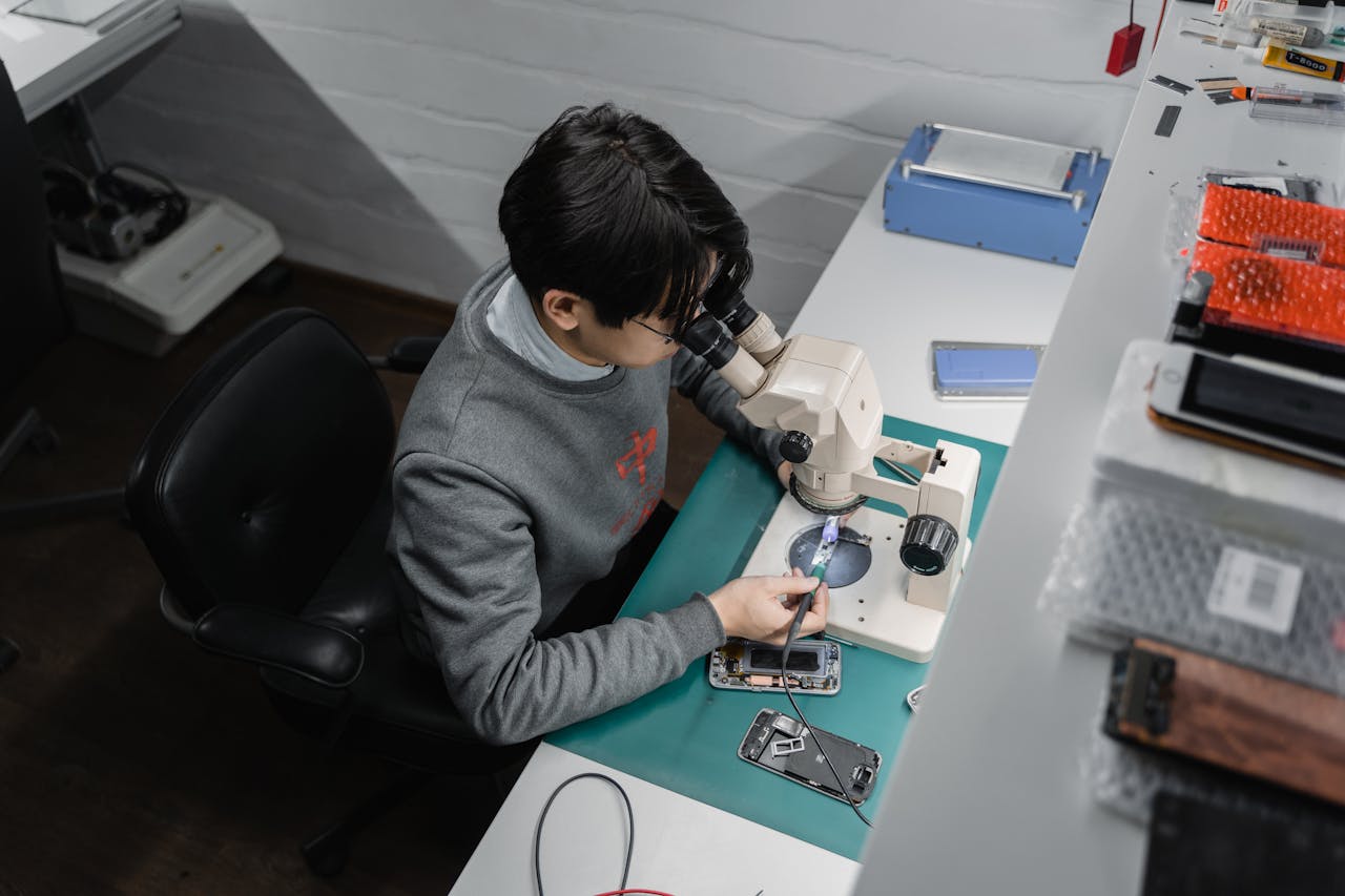 Home Technician works on smartphone repair under a microscope, indoors at a tech bench.